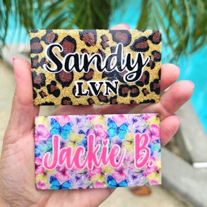 May include: Two rectangular name tags are displayed. The top tag has a leopard print background with the name "Sandy" in white script and "LVN" in black. The bottom tag features a butterfly pattern with the name "Jackie B." in pink script.