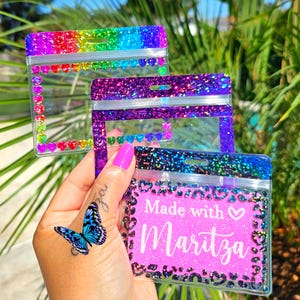 May include: Three clear badge holders with glitter and heart accents. One has a rainbow glitter top and heart border. Another has purple glitter and hearts. The third has a pink background with the text "Made with Maritza".