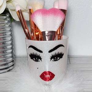 May include: White glitter makeup brush holder with a black and white face design, red lips, and long eyelashes. The holder is filled with various makeup brushes with pink and white bristles.