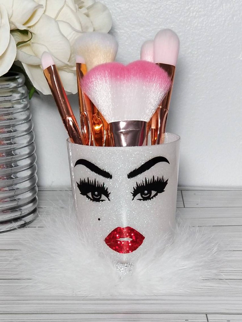 May include: White glitter makeup brush holder with a black and white face design, red lips, and long eyelashes. The holder is filled with various makeup brushes with pink and white bristles.