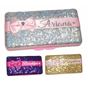 May include: Three sequin makeup bags with pink bows and personalized names. The bags are silver, purple, and gold.