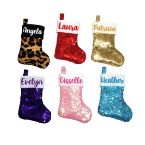 May include: Six Christmas stockings featuring sequined bodies in leopard print, red, gold, purple, pink, and blue. Each stocking has a white cuff with a personalized name printed in a script font: Angela, Laura, Patricia, Evelyn, Gisselle, and Heather.
