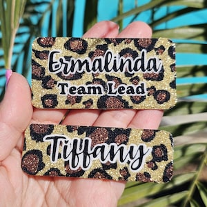 May include: Two gold and brown leopard print name tags. The top tag reads "Ermalinda Team Lead" and the bottom tag reads "Tiffany".