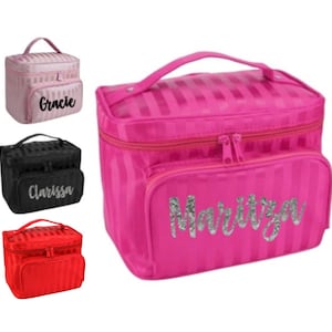 May include: A set of four cosmetic bags in pink, black, and red, each with a vertical striped pattern. The pink bag has the name "Maritza" in silver glitter, while the other bags have names "Gracie" and "Clarissa". Each bag has a top handle and a front pocket.