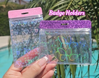 May include: Two clear plastic badge holders with glittery tops. One vertical holder features a pink glitter top and holographic heart patterns. The other horizontal holder has a purple glitter top and holographic butterfly designs. Text reads Badge Holders.