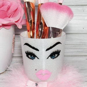 Pu&ograve; includere: Un portapennelli per il trucco bianco con glitter con un design di viso rosa e bianco. Il portapennelli ha un fiocco rosa e piume rosa intorno alla parte inferiore. Il portapennelli &egrave; pieno di pennelli per il trucco.