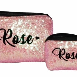 May include: Two pink sequined cosmetic bags with black zippers. The larger bag has a rectangular shape, and the smaller bag is rounded. Both bags have the name "Rose" in black script.