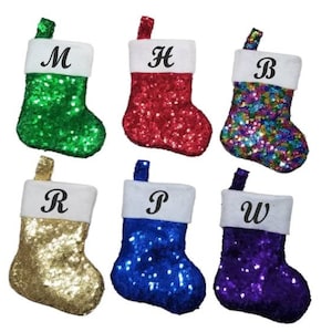 May include: Six Christmas stockings covered in shiny sequins in green, red, multicolored, gold, blue, and purple. Each stocking features a white cuff with a black letter printed on it, specifically M, H, B, R, P, and W, and a small hanging loop.