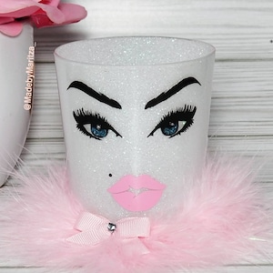 Pu&ograve; includere: Portapennelli per il trucco bianco con glitter, con un bordo di pelliccia rosa, un fiocco rosa con un strass e un'illustrazione in bianco e nero di un viso con occhi azzurri e labbra rosa. Il testo "@MadebyMaritza" &egrave; sul lato del portapennelli.