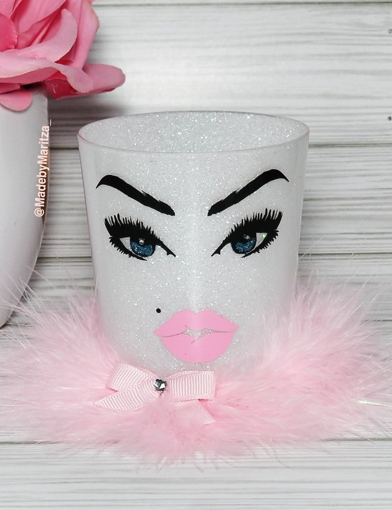 Pu&ograve; includere: Portapennelli per il trucco bianco con glitter, con un bordo di pelliccia rosa, un fiocco rosa con un strass e un'illustrazione in bianco e nero di un viso con occhi azzurri e labbra rosa. Il testo "@MadebyMaritza" &egrave; sul lato del portapennelli.