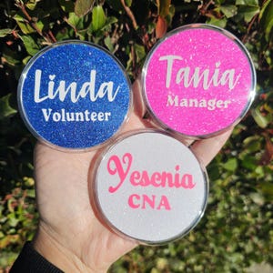 May include: Three round, glittery name badges in blue, pink, and white. The blue badge says "Linda Volunteer" in white script. The pink badge says "Tania Manager" in white. The white badge says "Yesenia CNA" in pink.