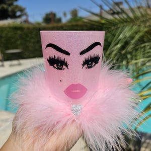 May include: Pink glitter tumbler with a black and white illustration of a face with eyelashes and lips. The tumbler is decorated with a pink feather boa and a rhinestone.