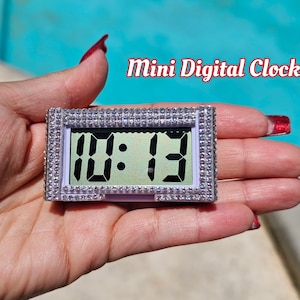 May include: A small white digital clock with a rhinestone border displays the time 10:13. The clock is being held in a hand with red nail polish.