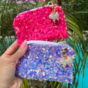 May include: Two rectangular pouches featuring a velvet base covered in shimmering sequins. The top pouch is bright pink, and the bottom pouch is iridescent blue. Each has a white zipper and a gold-tone clasp with a white bow charm attached.