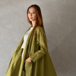 以下が含まれることがあります： 大きなポケットが付いた、オリーブグリーンのリネンコート。コートは開いており、白いトップスとパンツが見えます。モデルがコートを着ており、背景はニュートラルグレーです。
