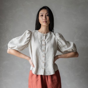 Puede incluir: Una mujer lleva una blusa de lino beige claro con mangas abullonadas y cierre de botones. También lleva pantalones de lino color óxido.