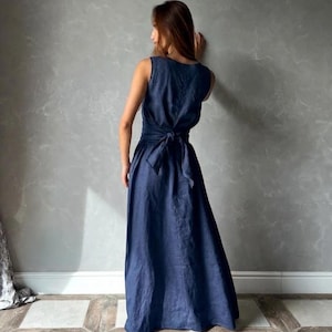 May include: A sleeveless, navy blue maxi dress with a tie-back detail. The dress has a flowing skirt and is made of a soft, draping fabric. The model is standing in front of a gray wall.