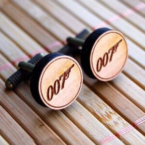 Op de afbeelding: Manchetknopen met een houten voorkant met het nummer "007" en een pistoolsilhouet. De manchetknopen hebben een zwarte rand en liggen op een lichtbruin houten oppervlak.