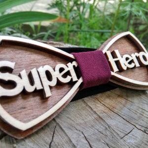 Puede incluir: Una pajarita de madera con las palabras "Super Hero" talladas en la madera. La pajarita tiene una correa ajustable de color burdeos.
