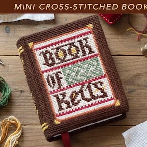 May include: A miniature book with a brown cross-stitched cover featuring the text "BOOK OF KELLS" in dark brown letters, accented with white, green, and red stitched patterns, and yellow corner details, with a red ribbon bookmark extending from the bottom.