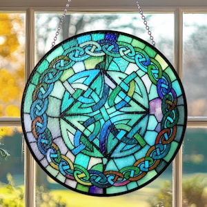 Puede incluir: Una vidriera redonda con un diseño de nudo celta. La ventana presenta un nudo central rodeado por una cadena de nudos en tonos de azul, verde y morado. La ventana está colgada frente a una ventana.