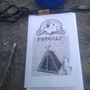 May include: A white booklet with the word "MAGUS" printed in black. The booklet has a black and white illustration of a tent and a person. A ball of blue yarn and a black cord with a copper accent are in the background.