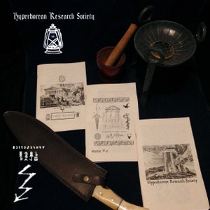以下が含まれることがあります： 黒い文字と画像が書かれた白い紙製の表紙の小さな本が3冊。本は「Mystae」、「Mystae V.2」、「Mystae VIII」というタイトル。本は、小さな茶色のすり鉢と乳棒、金属製の香炉、革で包まれたナイフのある黒い表面の上に置かれている。