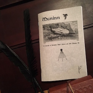 May include: A journal titled "Muninn" with a black and white illustration of a boat on water. The cover also features a raven, a starburst design, and text that reads "A Journal of Germanic Myth, History and Folk Memory". A black feather quill is visible.