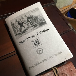 May include: A tan-colored book with the title "Hyperborean Pedagogy" printed in black. The book features a black and white illustration of people in a landscape and a black and white symbol of a sun with rays.