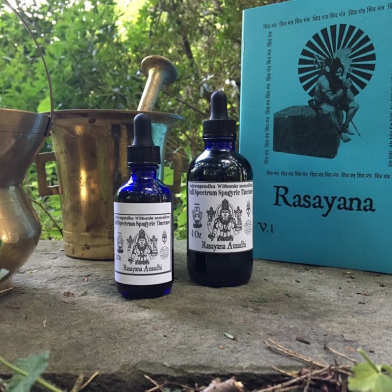 May include: Two blue glass bottles with dropper tops, labelled "Lavagandha Withania somnifera Full Spectrum Spagyric Tincture 1 oz Rasayana Ausadhi" and "Rasayana Ausadhi". The bottles are sitting on a grey stone surface with a blue book titled "Rasayana V.1" in the background.