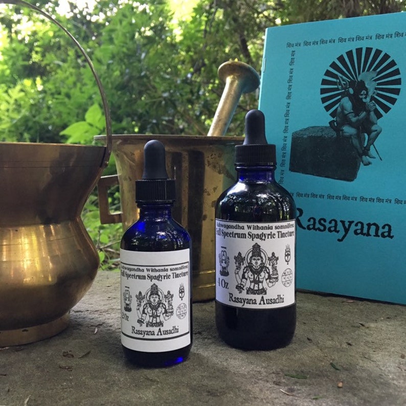 May include: Two small blue glass bottles with droppers, labelled "Withania somnifera Spectrum Spagyric Tincture" and "Rasayana Ausadhi". The bottles are sitting on a grey surface next to a book with a blue cover and the word "Rasayana" printed in white.