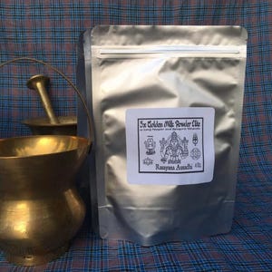 May include: A silver-colored stand-up pouch containing 40 oz of 5x Golden Milk Powder Elite with Long Pepper and Synergic Minerals, featuring a label with a deity illustration, positioned next to a brass mortar and pestle set.