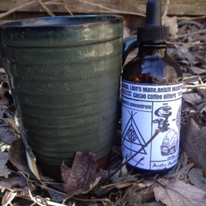 Puede incluir: Una taza de cerámica verde con base marrón junto a una botella de vidrio marrón con cuentagotas negro. La botella tiene una etiqueta blanca con texto e ilustraciones, incluyendo "Lion's Mane, Reishi Mushroom, cacao coffee bitters". La escena es al aire libre.