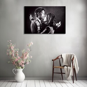 May include: Black and white artwork depicting a couple embracing, each holding a smartphone. The image is a grayscale painting on a wall, with a vase of pink flowers and a wooden chair in the foreground.