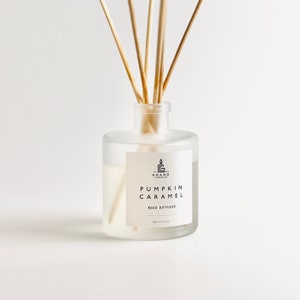 Może przedstawiać: Przezroczysty szklany dyfuzor patyczkowy z drewnianymi patyczkami. Etykieta głosi "Pumpkin Caramel Reed Diffuser" i zawiera logo świecy. Dyfuzor zawiera jasno zabarwioną ciecz i jest oznaczony "Net Wt 4 oz."