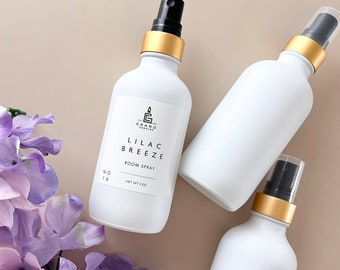 May include: Three white glass bottles with gold lids and black spray tops. The center bottle has a label that reads "Grand Candles" and "Lilac Breeze Room Spray" with a net weight of 4 oz.