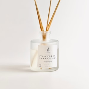 Puede incluir: Un difusor de varillas de vidrio transparente con varillas de madera. La etiqueta dice "Strawberry Cheesecake" y "Reed Diffuser". La botella contiene un líquido de color claro y está etiquetada como "Net wt 4 oz".