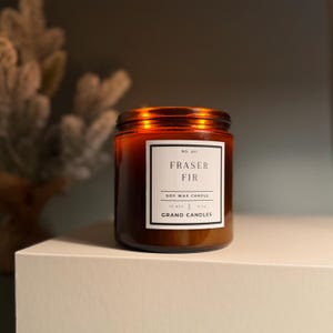 May include: A brown glass jar candle with a white label that reads "FRASER FIR" and "SOY WAX CANDLE". The candle is 454g and has a burn time of 70 hours. The brand name "GRAND CANDLES" is also printed on the label.