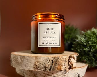 Könnte beinhalten: Eine braune Glas-Kerze mit kupferfarbenem Deckel, beschriftet mit "Blue Spruce" auf einem weißen Etikett. Die Kerze steht auf einer Holzscheibe, mit Grünzeug im Hintergrund. Die Kerze ist eine Sojawachskerze.