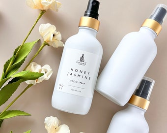 May include: Three white frosted glass bottles with gold lids and black spray tops. The bottles are labeled "The Grand Candles" and "Honey Jasmine Room Spray" with a net weight of 4 oz. The bottles are on a light brown surface with white flowers.