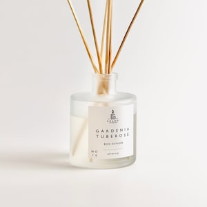 May include: A clear glass bottle with a white label that reads "GARDENIA TUBEROSE REED DIFFUSER NO 13 NET WT 4 OZ" and a small logo of a candle with the letter "G" inside. The bottle is filled with a clear liquid and has wooden sticks sticking out of the top.
