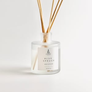 Puede incluir: Un difusor de varillas de vidrio transparente con varillas de madera. La etiqueta dice "Blue Spruce Reed Diffuser" y "Net Wt 4 oz". La botella tiene un aspecto esmerilado y está llena de líquido transparente.