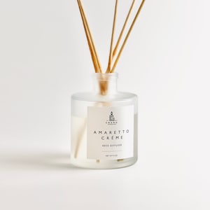 Puede incluir: Un difusor de caña de vidrio transparente con una etiqueta blanca que dice "AMARETTO CRÉME REED DIFFUSER" y "NET WT 4 OZ." La botella contiene un líquido de color claro y varias cañas de madera.