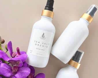 Op de afbeelding: Drie witte glazen flessen met gouden deksels en zwarte spuitkoppen. De fles in het midden heeft een etiket met de tekst "Sea Salt Orchid Room Spray" en "Net Wt 4 oz".