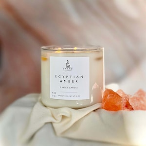May include: A clear glass candle jar with a white label that reads "EGYPTIAN AMBER 3 WICK CANDLE." The candle is filled with a creamy wax and sits on a white cloth. Orange crystals are to the right of the candle.