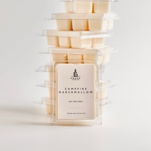 May include: A stack of soy wax melts in clear plastic packaging. The label reads "Campfire Marshmallow" and "Soy Wax Melt." The wax melts are a light beige color, and the packaging is stacked on a white background. The brand name is "Grand Candles."