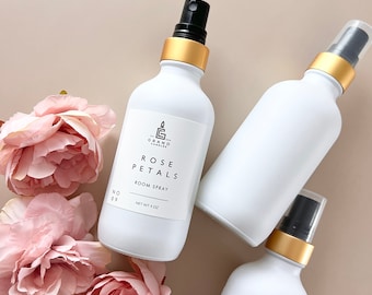 Op de afbeelding: Drie witte glazen flessen met gouden deksels en zwarte spuitkoppen. De middelste fles heeft een etiket met de tekst "The Grand Candle Co. Rose Petals Room Spray Net Wt 4 oz."