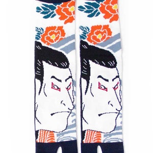 Op de afbeelding: Een paar witte tabi-sokken met een Japans geïnspireerd ontwerp in blauw, rood en oranje. De sokken tonen het gezicht van een traditionele Japanse man met een strenge uitdrukking. De sokken hebben een gespleten teenontwerp.