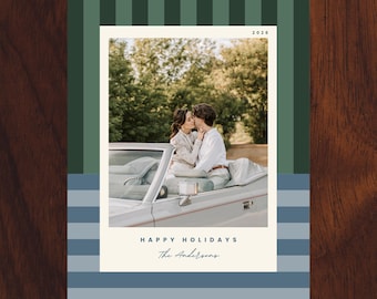 May include: A rectangular holiday card featuring a central photograph of two people in a convertible car. The top border has dark green vertical stripes, the bottom has blue horizontal stripes. Text reads 2026, HAPPY HOLIDAYS, and The Andersons.