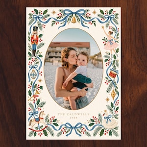 May include: A rectangular holiday photo card featuring a central oval frame containing a photograph of a person holding a child on a beach. The border is decorated with illustrations of a nutcracker, ballerina, swan, and rocking horse with blue ribbons.